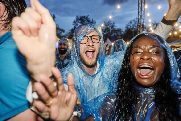 Festival-goers dance and sing in the rain, wearing blue ponchos.