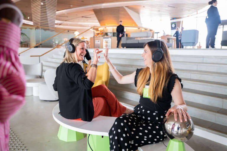 Two smiling participants wearing headphones interact in a bright and modern welcome area.